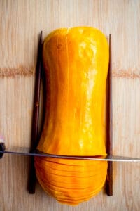 half of a honeynut squash on a wooden cutting board with a chopstick on either side and a knife slicing the squash crosswise