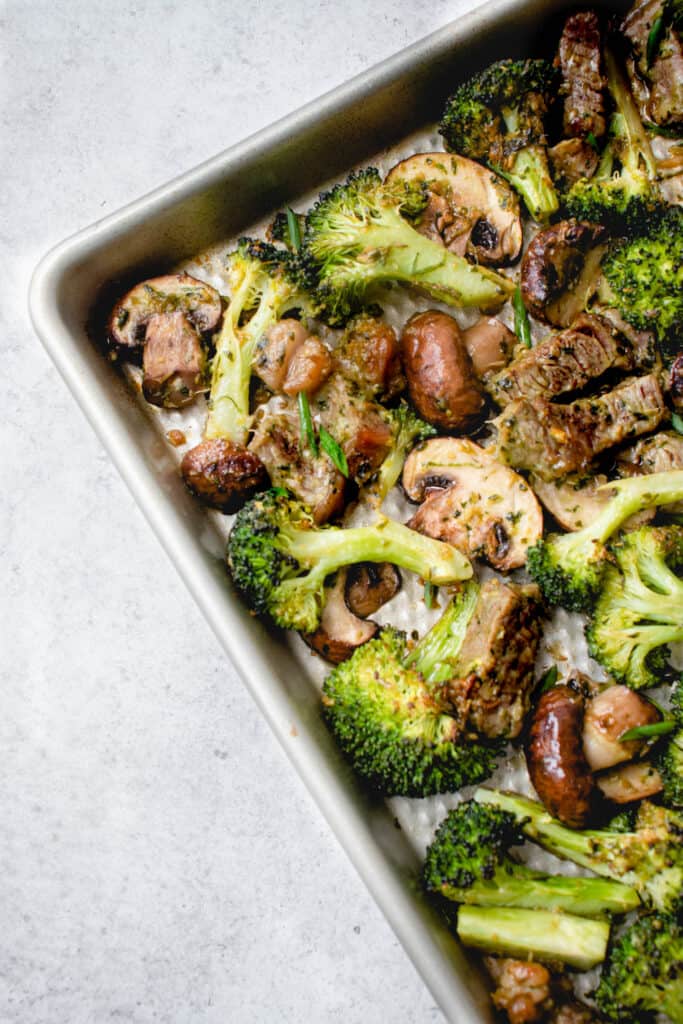 stainless steel sheet pan with beef, broccoli and mushrooms at an angle against a speckled grey background