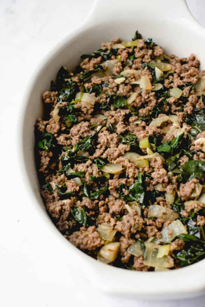 white oval casserole dish with ground beef, onions, celery and kale