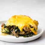 one slice of beef and kale casserole topped with cheese sauce on a small white plate against a grey background and white wall