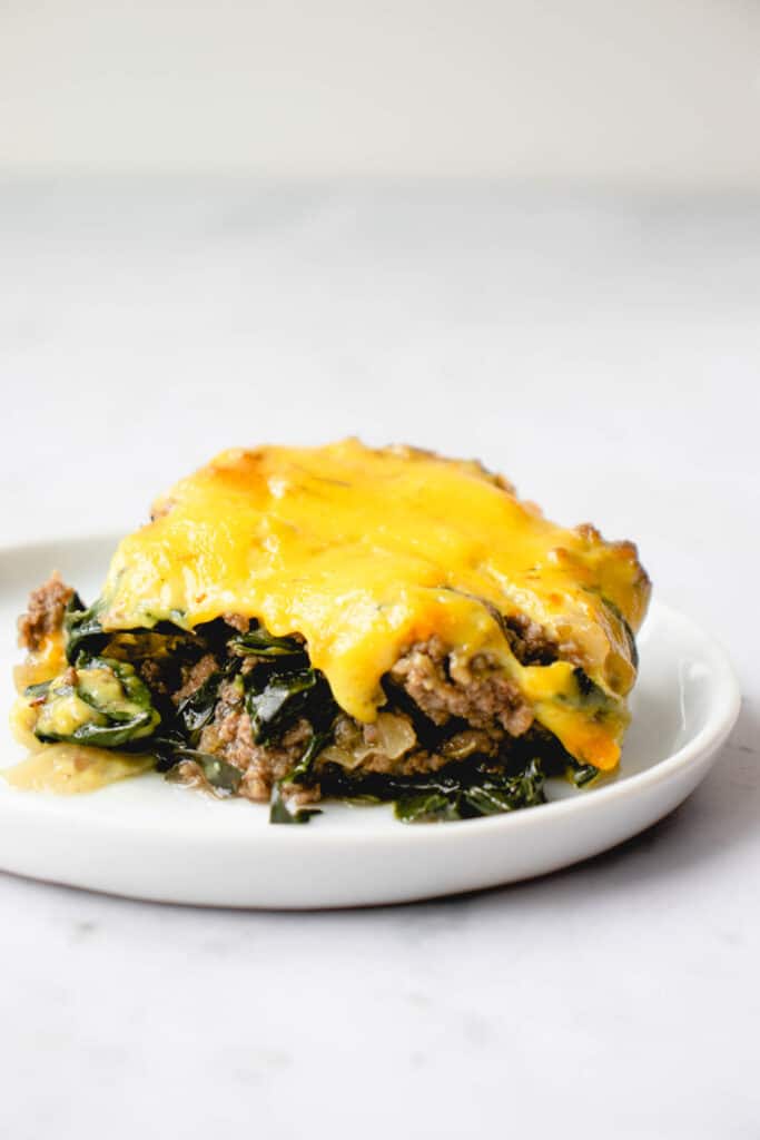 one slice of beef and kale casserole topped with cheese sauce on a small white plate against a grey background and white wall 