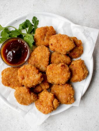 white plate with parchment paper with chicken nuggets and a glass bowl with a brown sweet and sour sauce and garnish of cilantro against a grey background