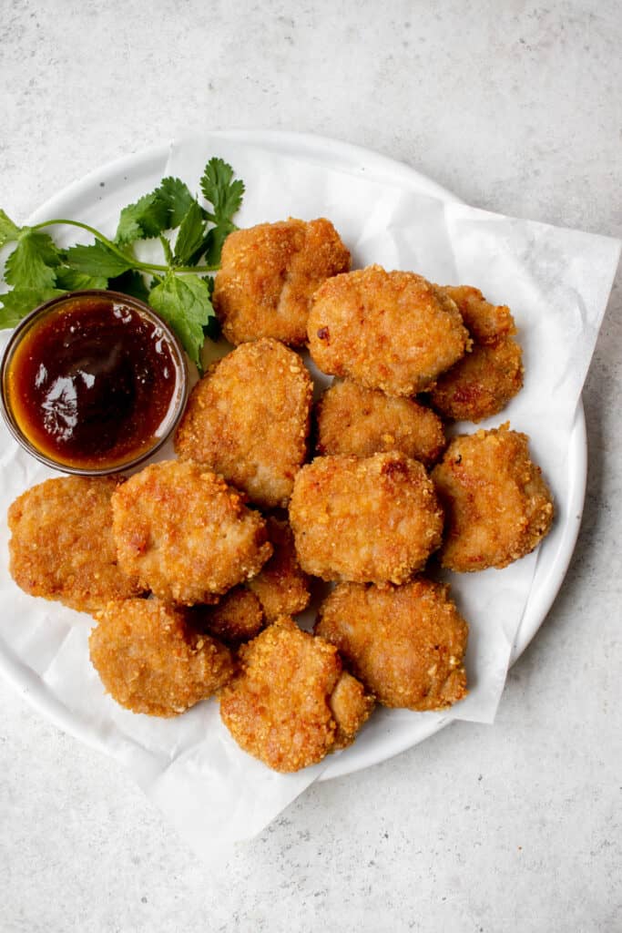 white plate with parchment paper with chicken nuggets and a glass bowl with a brown sweet and sour sauce and garnish of cilantro against a grey background