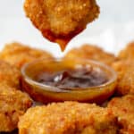 hand dipping a chicken nugget into a small glass bowl with brown sweet and sour sauce surrounded by chicken nuggets on a piece of parchment paper