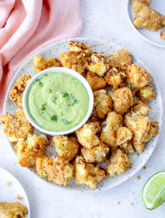 grey speckled plate with air fryer cauliflower bites surrounding a small white bowl with green cilantro lime sauce with two small white plates with crispy cauliflower florets, a lime wedge and a light pink cloth napkin in the top left corner