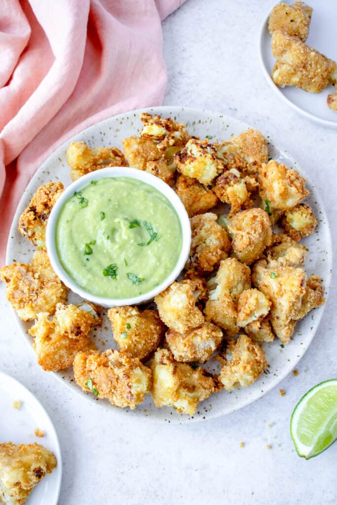 grey speckled plate with air fryer cauliflower bites surrounding a small white bowl with green cilantro lime sauce with two small white plates with crispy cauliflower florets, a lime wedge and a light pink cloth napkin in the top left corner