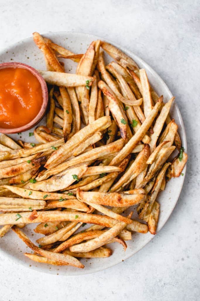 light beige speckled plate with green banana fries and a small wooden bowl with carrot ketchup against a light grey backdrop