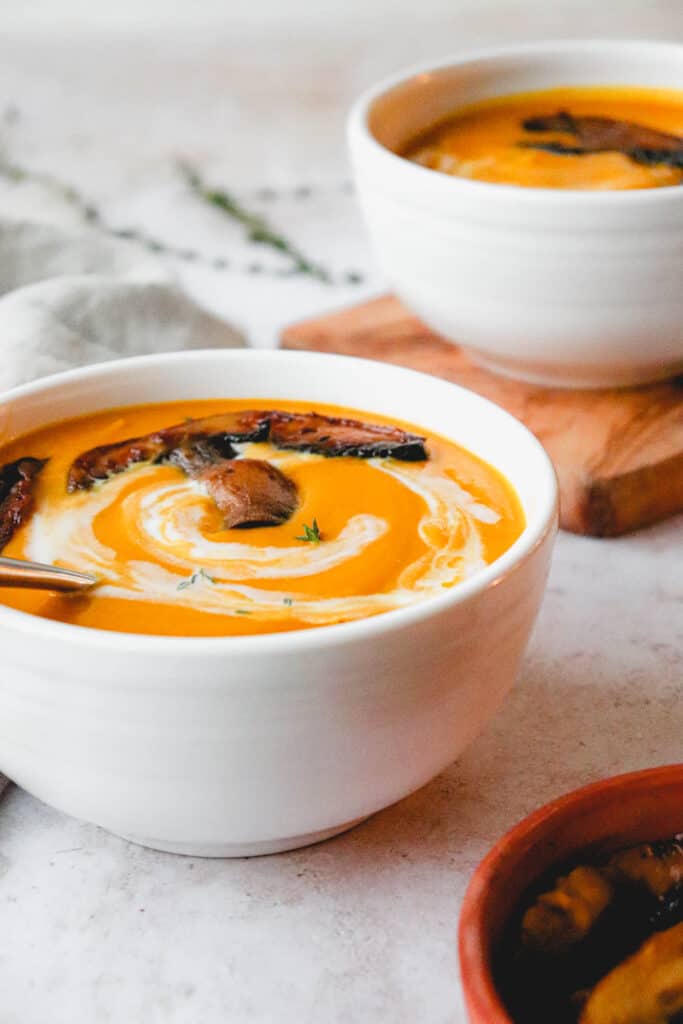 side shot of two white bowls with roasted squash soup, with sliced portobello mushroom garnish and coconut milk swirl on a light background with a beige cloth napkin, sprigs of fresh thyme and a and a clay plate with sliced portobello mushrooms