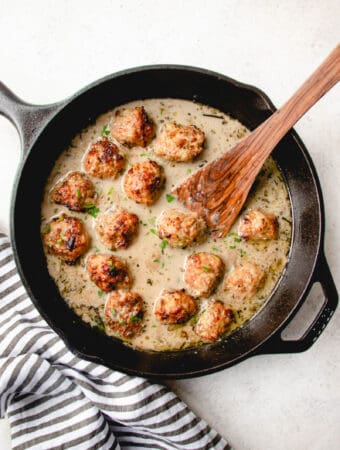 black cast iron skillet with chicken meatballs in cream sauce with wooden spoon on a grey background with a grey and white cloth napkin on the bottom left