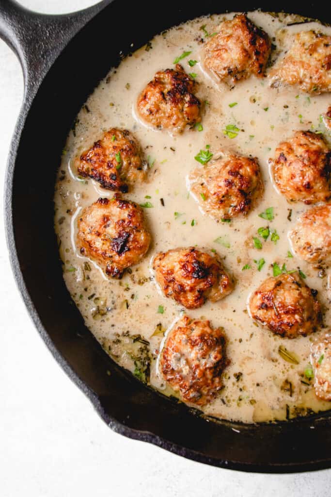 black cast iron skillet with chicken meatballs in a cream sauce, garnished with chopped parsley on a white background