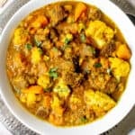 wide white bowl with lamb curry, sweet potatoes and cauliflower, garnished with chopped cilantro on a silver platter