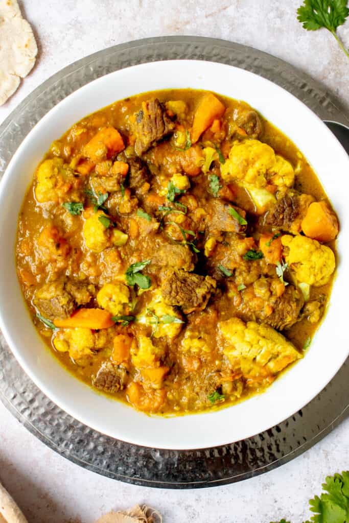 wide white bowl with lamb curry, sweet potatoes and cauliflower, garnished with chopped cilantro on a silver platter 