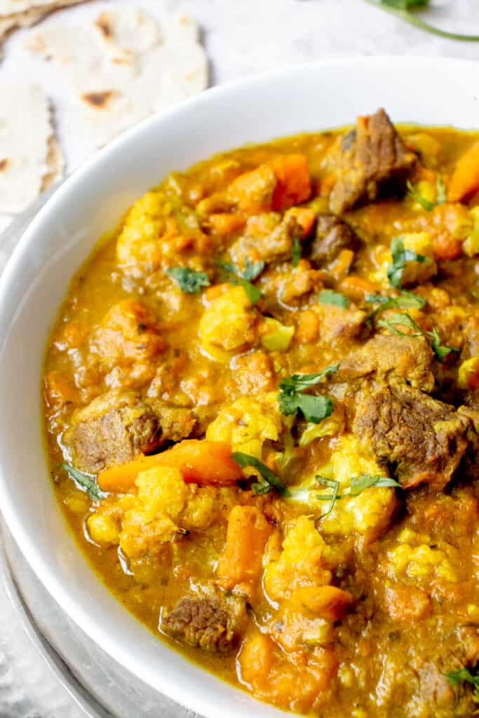 wide white bowl with lamb curry, sweet potatoes and cauliflower, garnished with chopped cilantro on a silver platter with a serving spoon on a light grey background with pieces of teared flatbread on the top lefthand corner of the frame