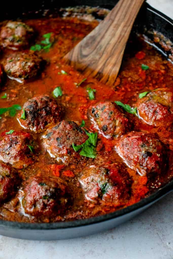 black cast iron skillet with lamb meatballs in nomato sauce with chopped parsley and a wooden spoon