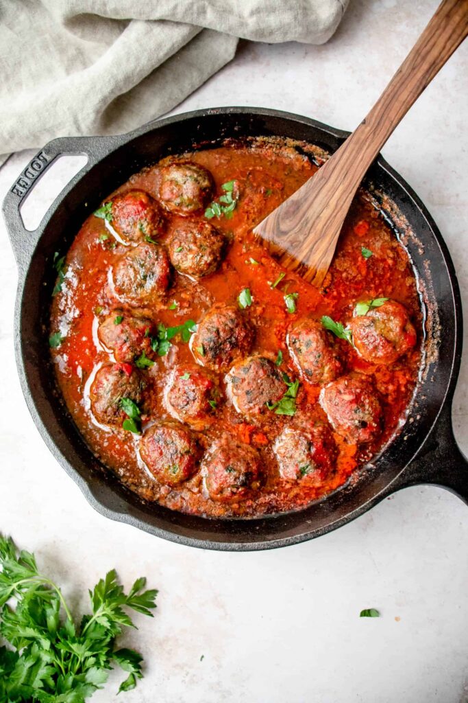 black cast iron skillet with lamb meatballs in nomato sauce with wooden spoon on a grey background with sprigs of cilantro on the bottom left and a beige cloth napkin on the top left