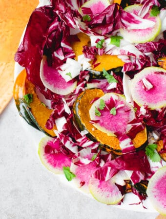 large white plate with salad with radicchio, watermelon radish, sliced acorn squash, chopped apple and chopped cilantro, with one wooden salad spoon to the top left