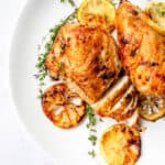 half of a white plate with two chicken breasts, one of which has been partially sliced, garnished with sliced lemons and sprigs of thyme