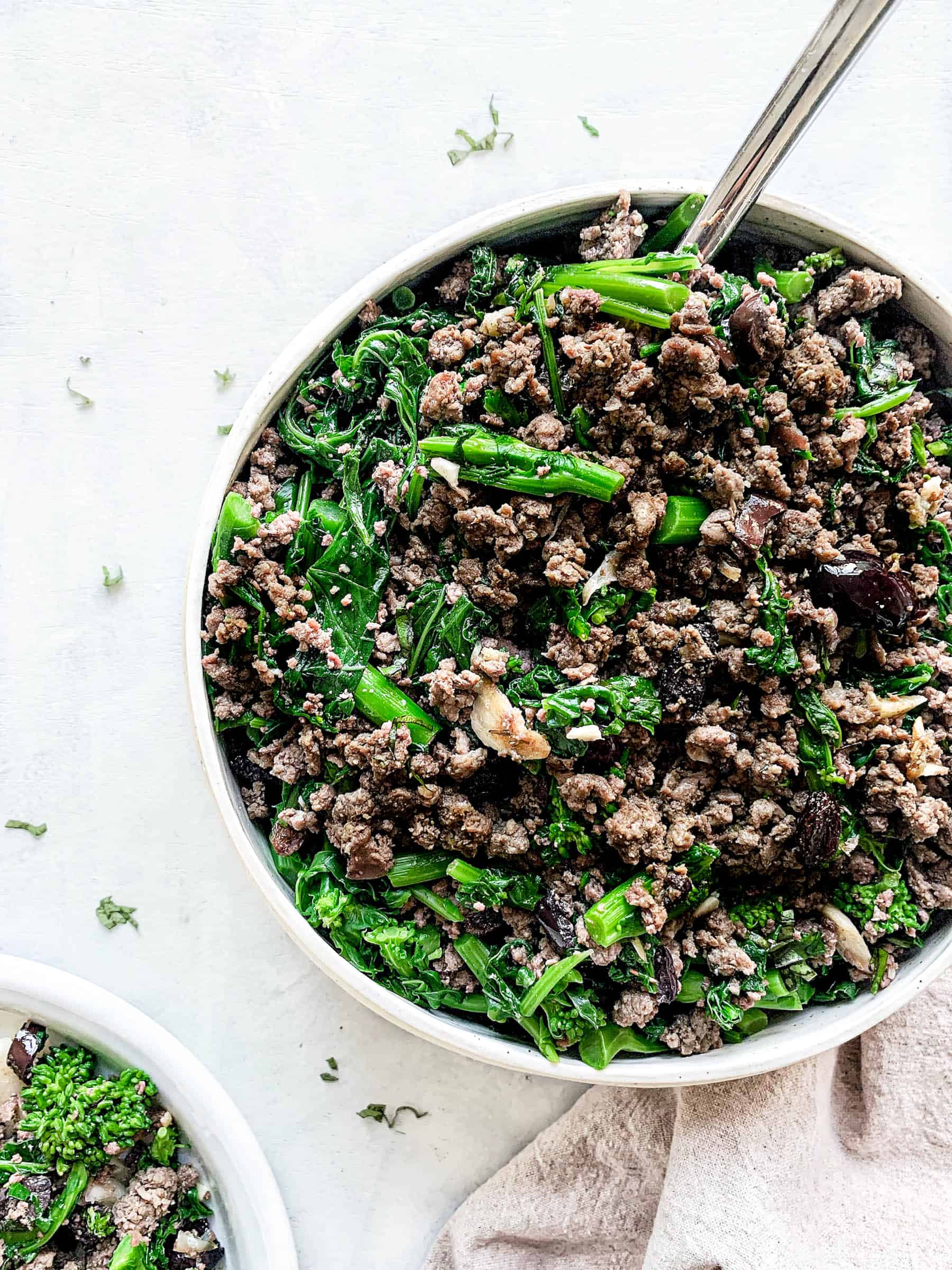 large round dish with ground beef, red olives, raisins and broccoli rabe with a spoon and a beige cloth napkin