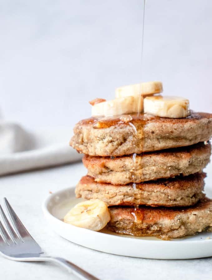 white plate with stack of four banana pancakes topped with sliced bananas, with a small fork to the left and a beige cloth napkin in the background with a drizzle of maple syrup