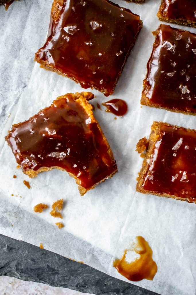 five caramel blondies with flaky sea salt on parchment paper on top a grey slate with one bite taken out of the bottom left blondie