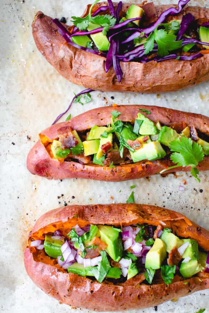 overhead shot of three baked sweet potatoes slit down the middle that are stuffed with purple cabbage, cubed avocado, bacon ad cilantro on a piece of light brown parchment paper with chopped cilantro and a lime wedge on the side