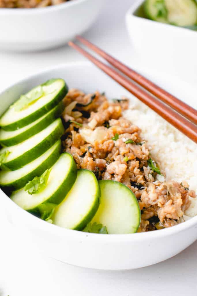 two white bowls with sliced cucumber, ground pork and finely chopped cauliflower with two brown chopsticks on one bowl and a small saucer with chopped cucumber in the far right corner 