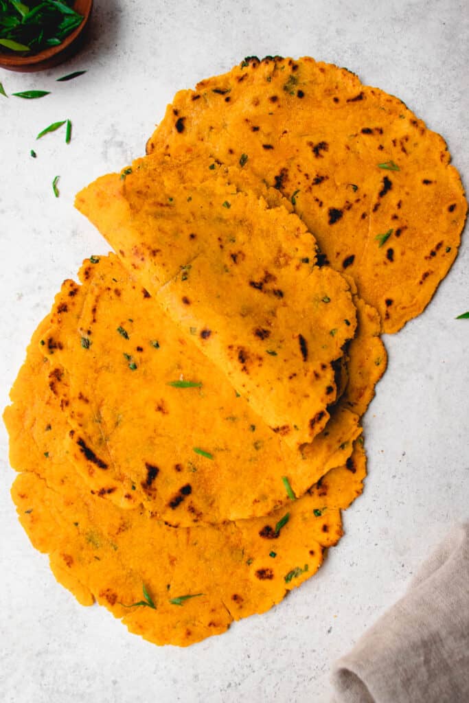 four sweet potato tortillas on a light grey background with one tortilla folded in half and garnished with chopped chives and a small wooden bowl in the top left corner with chopped chives and a brown cloth napkin on the bottom right