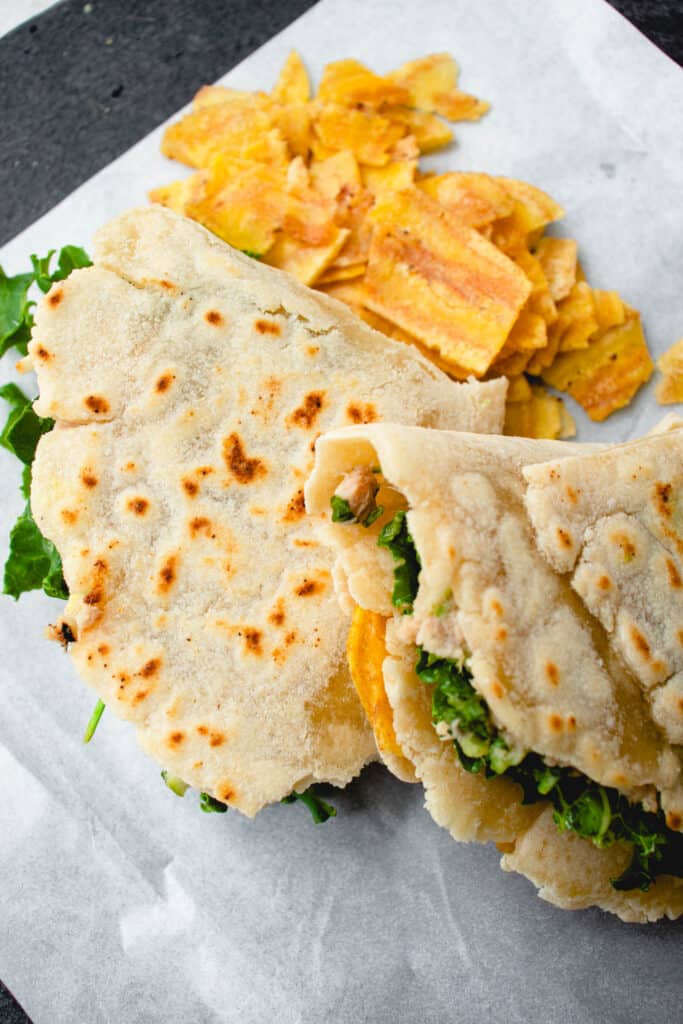 two cassava flour wraps stuffed with tuna salad, kale, avocado and plantain chips on a piece of parchment paper with plantain chips
