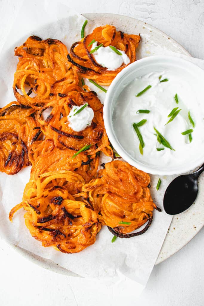 light beige speckled plate with a sheet of white parchment paper with butternut squash latkes, topped with sour cream and chives a small white bowl with sour cream and chives and a small black spoon to the right of the bowl