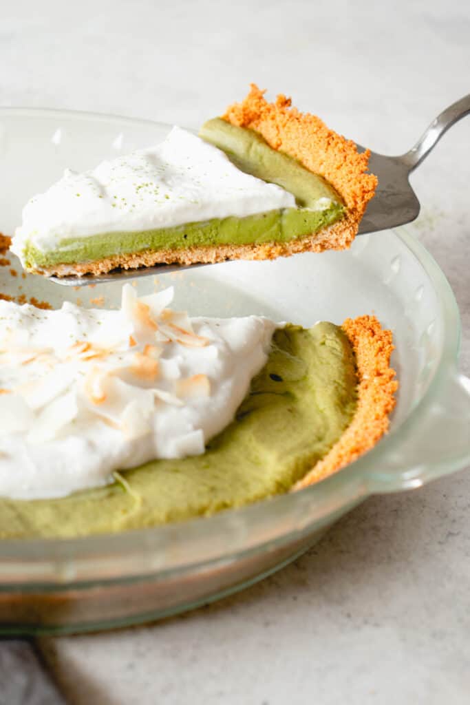 glass pie dish against a grey background with green matcha pie topped with whipped coconut cream with a stainless steel pie knife lifting one slice up