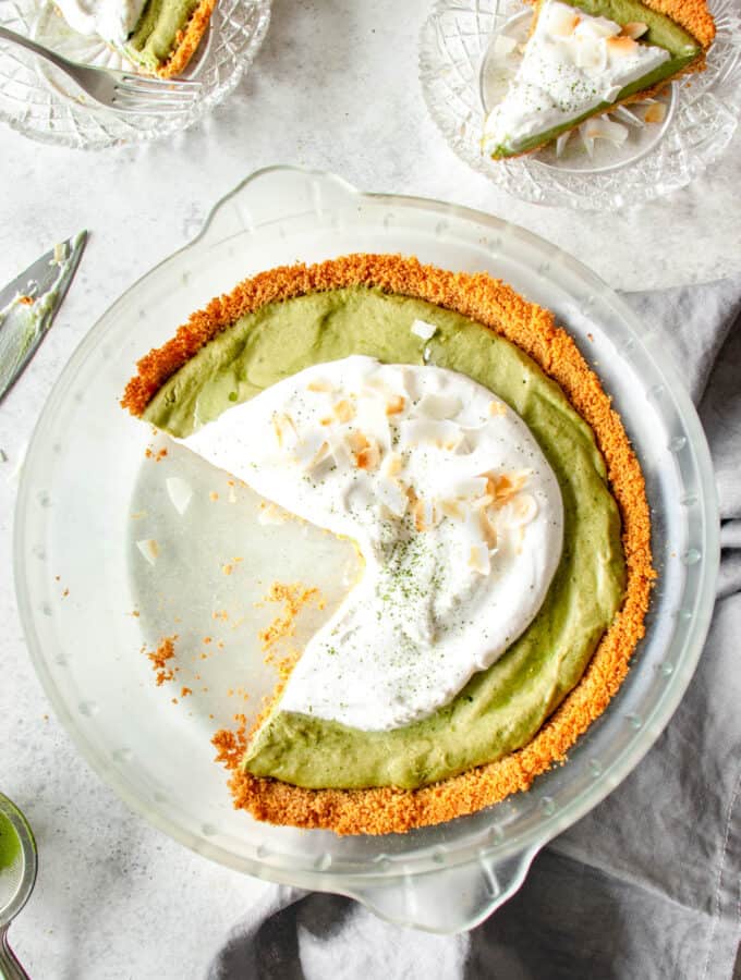 glass pie dish against a grey background with green matcha pie topped with whipped coconut cream and toasted coconut flakes with two glass pie plates with one slice of pie in each and a dark grey cloth napkin on the right side of the pie plate