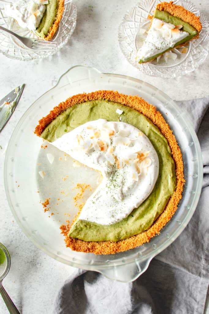 glass pie dish against a grey background with green matcha pie topped with whipped coconut cream and toasted coconut flakes with two glass pie plates with one slice of pie in each and a dark grey cloth napkin on the right side of the pie plate