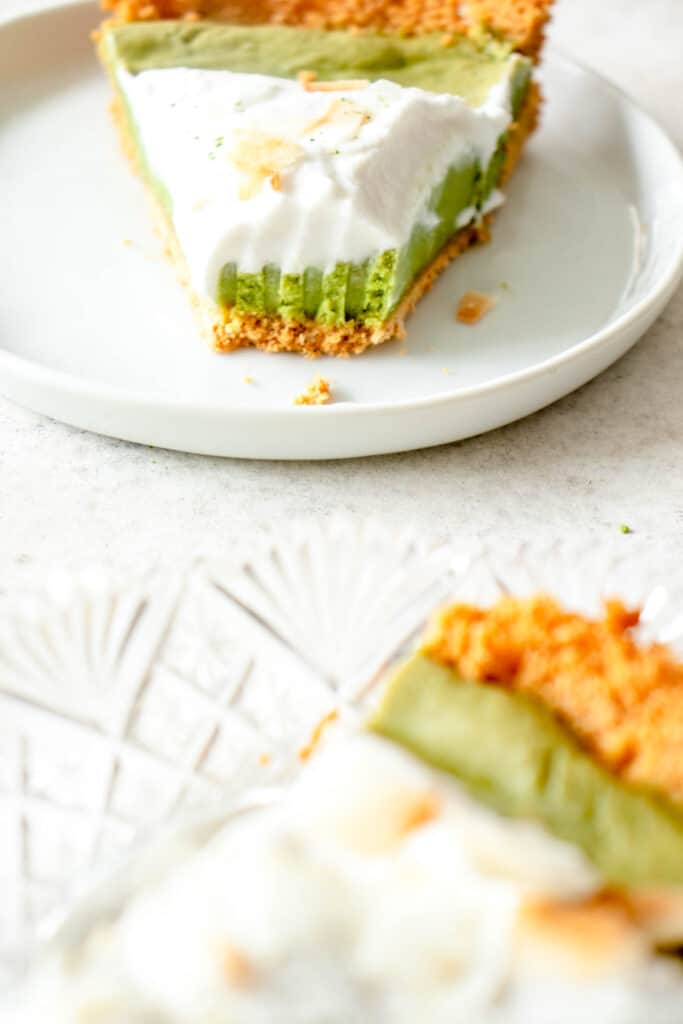 one glass plate with a slice of coconut matcha pie in the foreground with a small white plate and another slice of the matcha pie in the background topped with whipped coconut cream with one bite eaten from the slice of pie revealing the texture