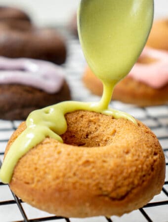 black cooling rack with pastel-glazed mini donuts in the background and a vanilla donut being drizzled with a bright green icing with a spoon in the foreground