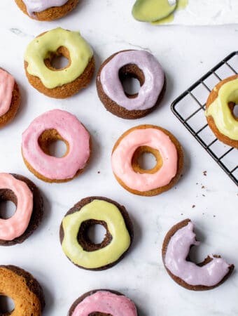 a series of lavender, pink and green frosted chocolate and vanilla mini donuts against a marble backdrop with a stainless steel spoon with green frosting on a white piece of parchment paper at the top right of the image and a corner of a black cooling rack with one glazed donut on the right side of the frame
