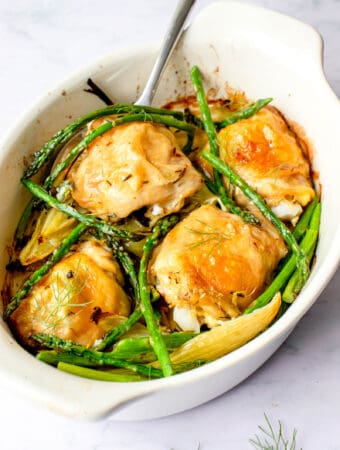 white oval shaped baking dish with four chicken thighs, asparagus and fennel with a stainless steel serving spoon against a white and grey marbled background