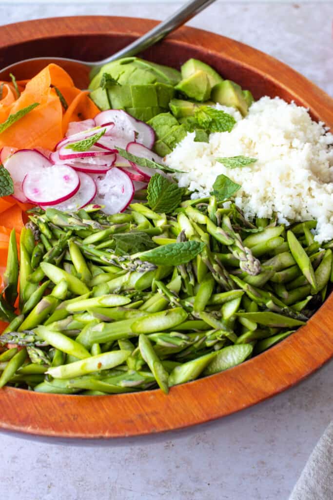 a large wooden bowl with sliced asparagus, shaved carrots, shaved radishes and riced cauliflower with a large serving spoon made of stainless steel sticking out the bowl placed on a light beige patterned backdrop