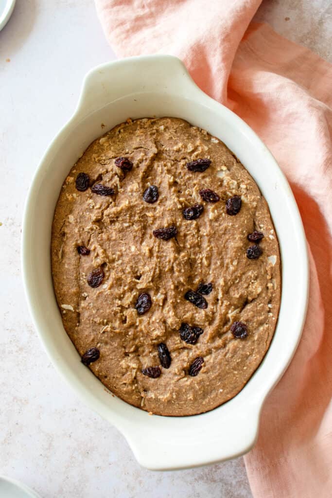 white oval shaped baking dish with baked oatmeal topped with raisins on a light brown speckled backdrop with a light pink cloth napkin gathered on the right side of the baking dish running vertically from the top to the bottom of the frame.