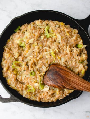 black cast iron pan on a grey and white marbled background filled with risotto made with orzo, leek and fennel with some slices of green leeks prominent on the surface and a wooden flat spoon dipped into the pan in the bottom right quarter