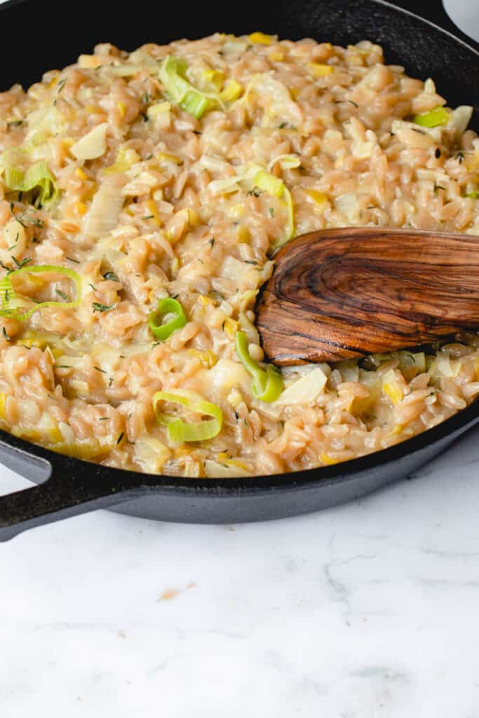black cast iron pan on a grey and white marbled background filled with risotto made with orzo, leek and fennel with some slices of green leeks prominent on the surface and a wooden flat spoon dipped into the pan in the bottom right corner of the frame