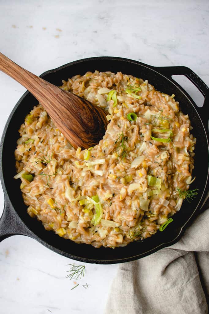 black cast iron pan on a grey and white marbled background filled with risotto made with orzo, leek and fennel with some slices of green leeks prominent on the surface and a wooden flat spoon dipped into the pan in the top left quarter and a light beige cloth napkin tucked under the pan in the bottom right corner 