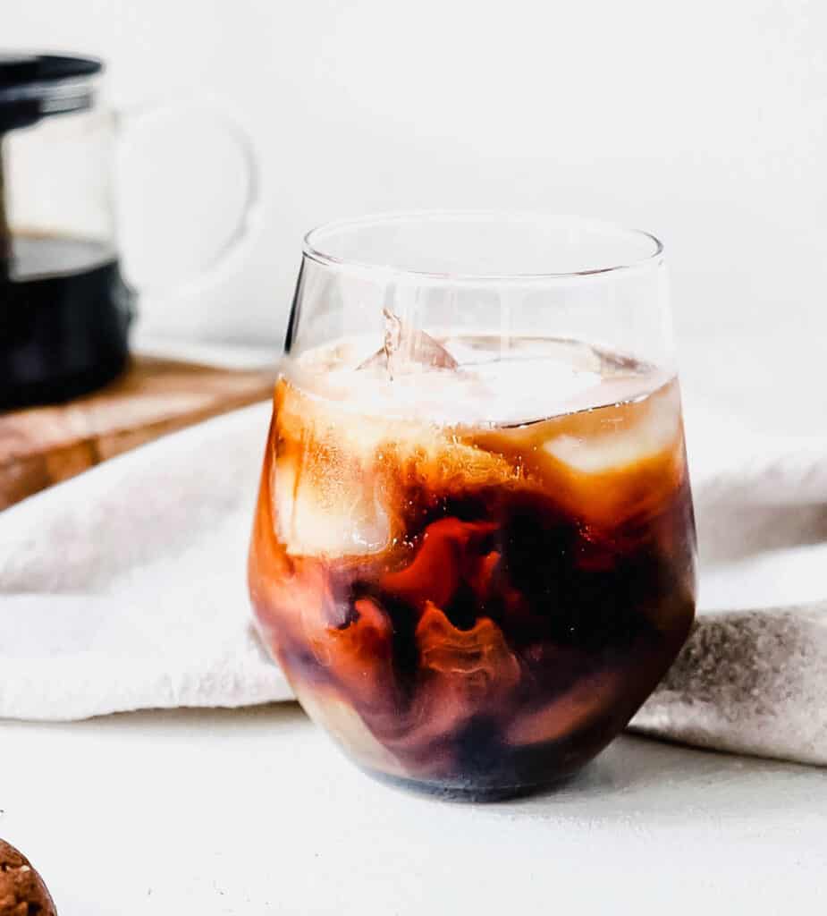 glass with dark brown dandelion chicory tea and coconut milk creating a swirled pattern against a white backdrop with a light beige cloth napkin behind the glass and a glass teapot with black tea sitting on top a small wooden cutting board in the background