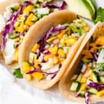 three soft tacos on a white piece of parchment paper filled with white fish, mango and cucumber salsa, red cabbage, and cilantro with sliced avocado on the side