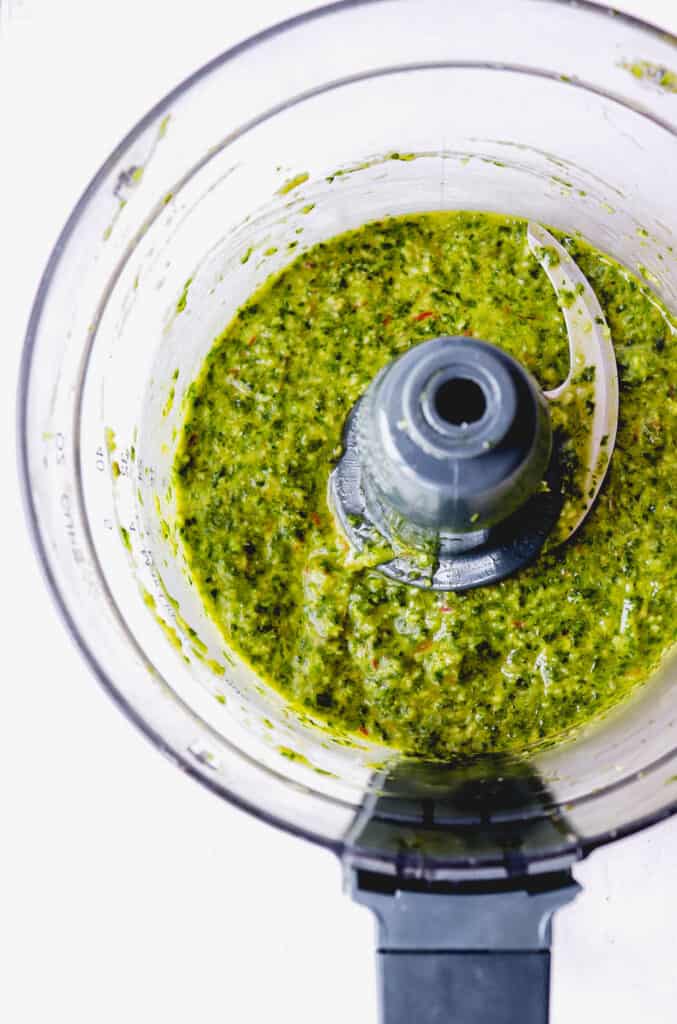 overhead shot of a food processor with a grey handle filled with pesto sitting on a white surface