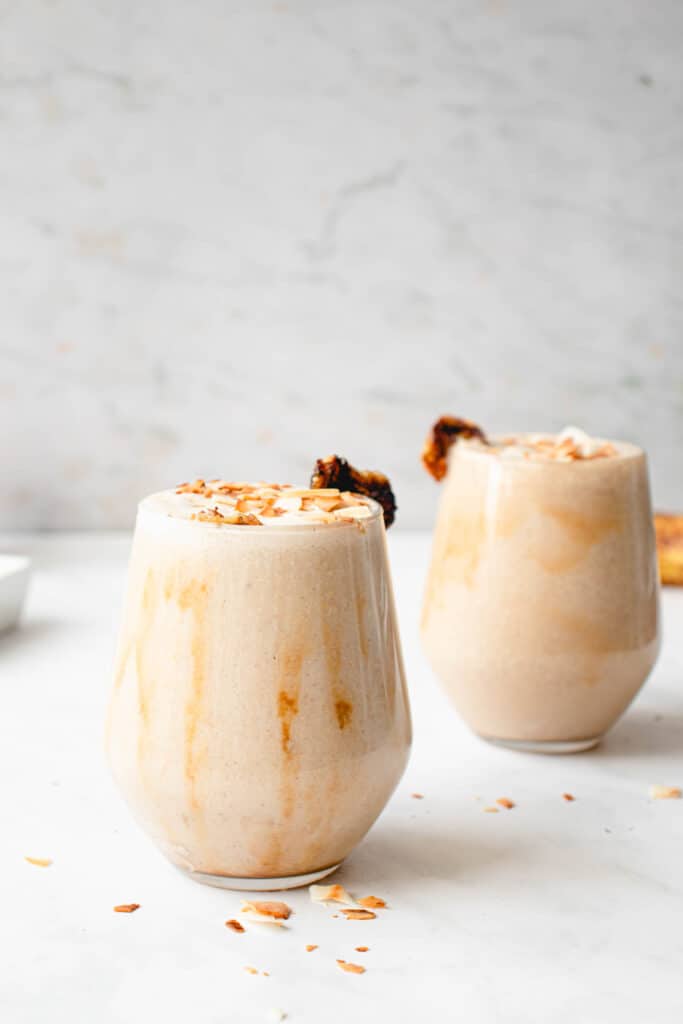 two glasses drizzled with caramel sauce on the inside and filled with banana milkshake.The milkshakes are topped with toasted coconut flakes and garnished with a slice of caramelized banana on the side of each of the glasses. They sit on a white marbled background and the backdrop is a darker grey marbled patter