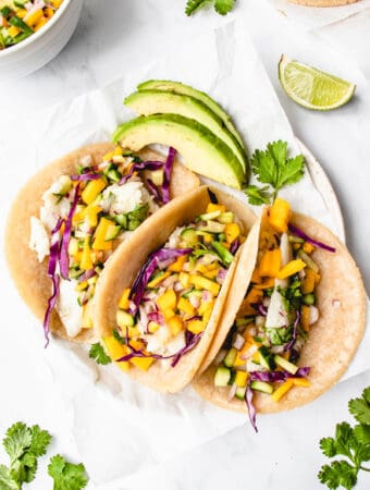 overhead shot of three soft tacos on a white piece of parchment paper filled with white fish, mango and cucumber salsa, red cabbage, and cilantro with sliced avocado on the side and lime wedges surrounding the tacos. In the top left corner, the side of a white bowl with mango salsa is visible and in the top right corner there are some fresh tortillas on a piece of parchment paper