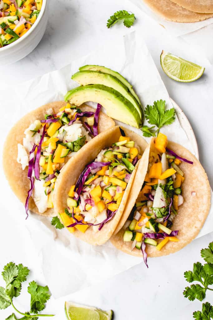 overhead shot of three soft tacos on a white piece of parchment paper filled with white fish, mango and cucumber salsa, red cabbage, and cilantro with sliced avocado on the side and lime wedges surrounding the tacos. In the top left corner, the side of a white bowl with mango salsa is visible and in the top right corner there are some fresh tortillas on a piece of parchment paper