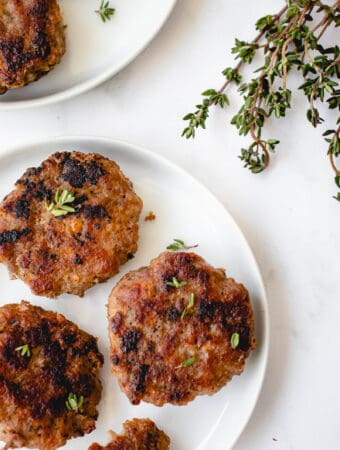Two small white plates with homemade pork breakfast sausage patties garnished with fresh thyme leaves. There is a bunch of sprigs of thyme in the top right of the image