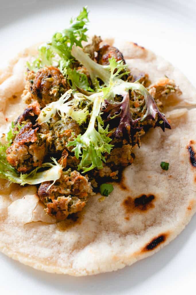a charred cassava flour tortilla sits on top a white plate with crushed pork meatballs, an herbed green sauce and sprigs of lettuce