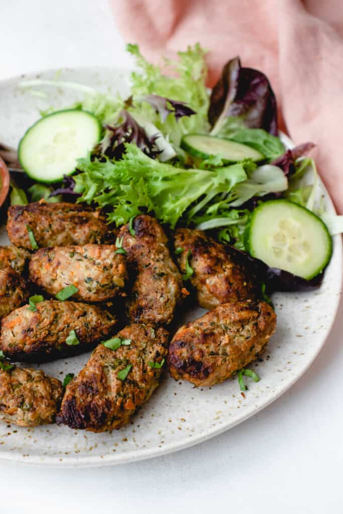 light brown speckled plate with pork meatballs stacked in a pile. o. The meatballs surround a small wooden bowl with an herbed lime sauce and a green salad with cucumbers